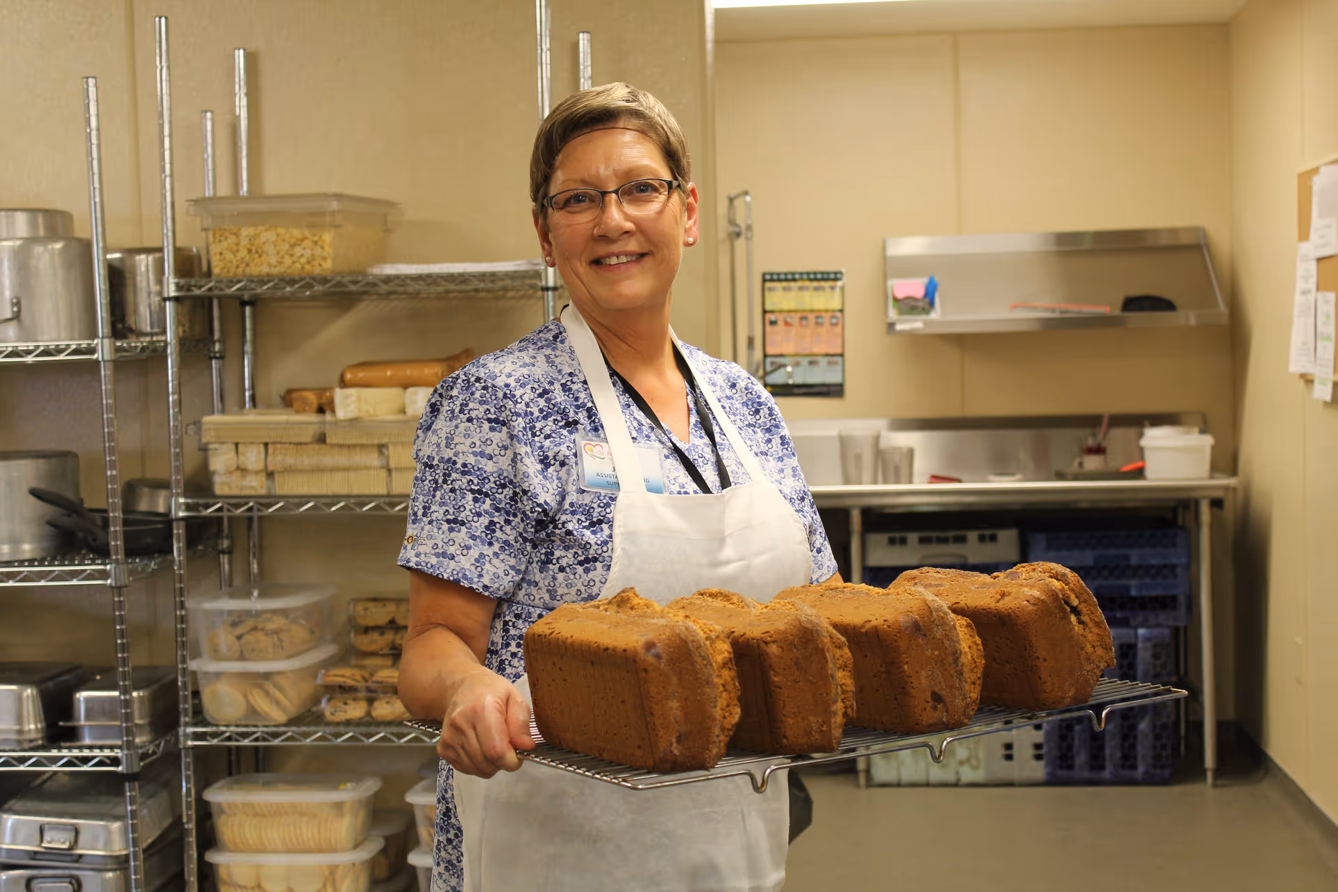 A smiling woman in an apron holding a cooling rack of freshly baked loaves in a commercial kitchen.