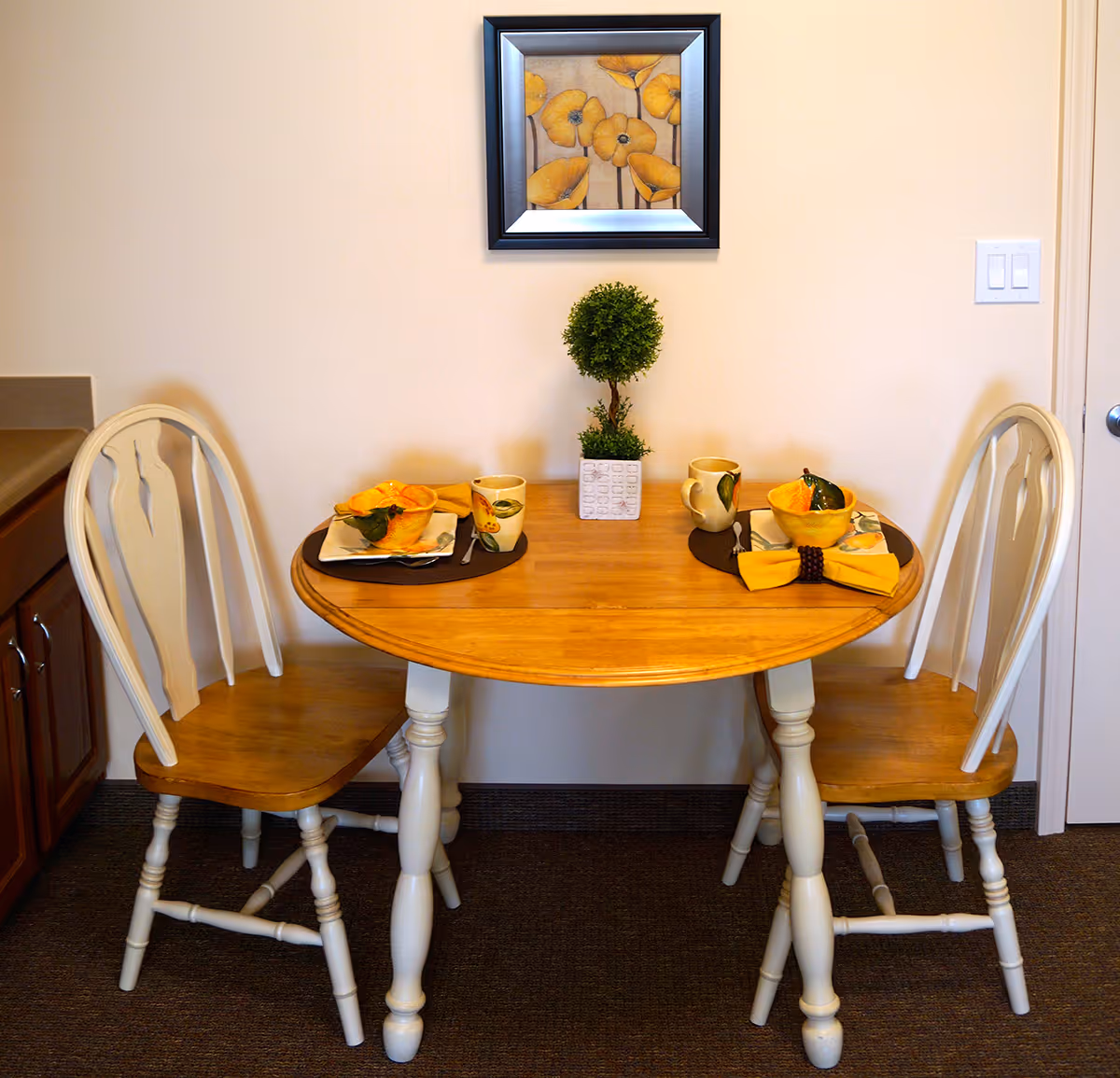 A small wooden dining table set for two with matching wooden chairs. The table has two place settings with yellow napkins, cups, bowls, and utensils. A small potted plant is centered on the table. A framed painting of yellow flowers hangs on the beige wall behind the table.