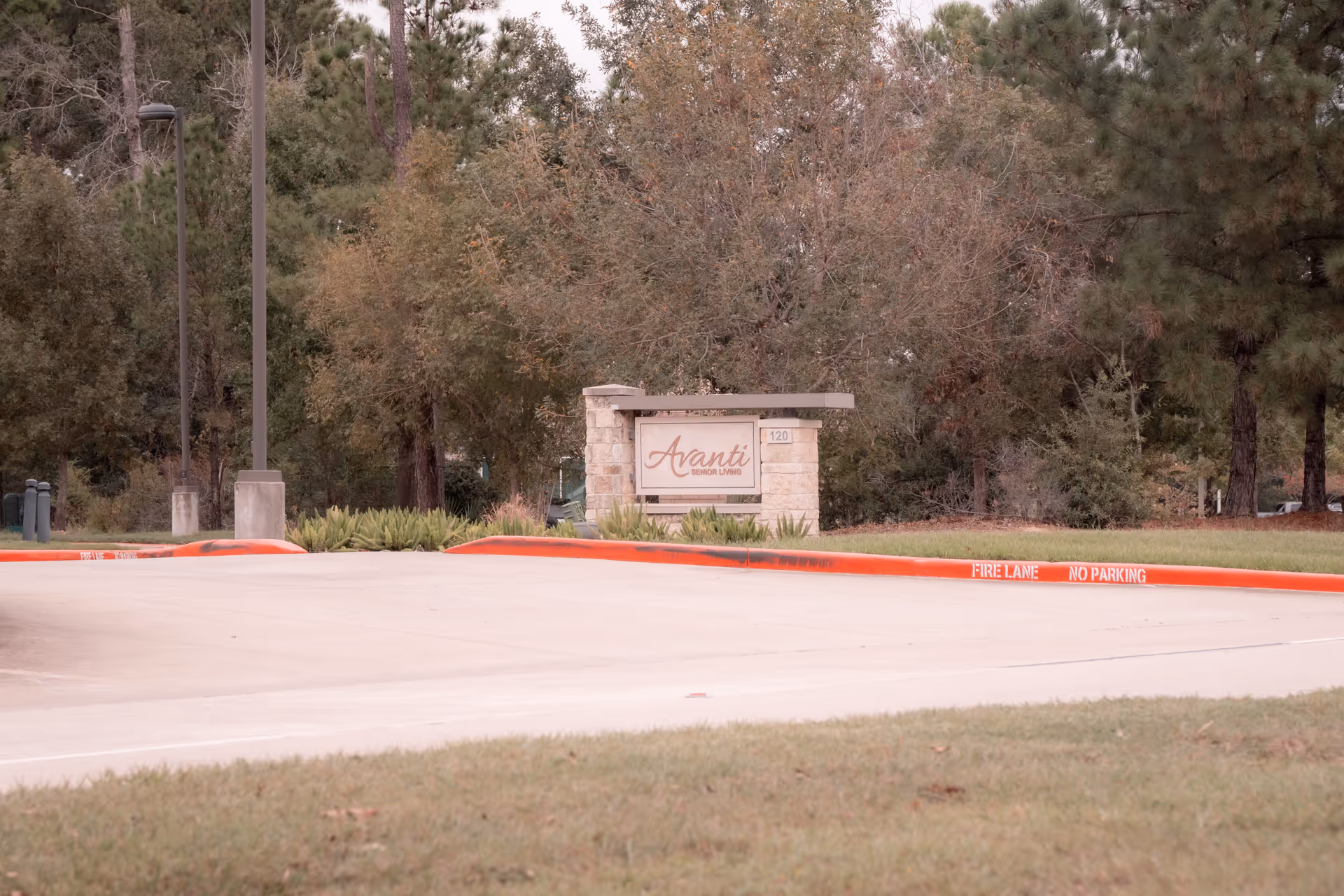 Stone sign for Avanti Senior Living at Vision Park surrounded by trees and greenery, with a concrete driveway and red-painted curb marked 'FIRE LANE NO PARKING' in the foreground.