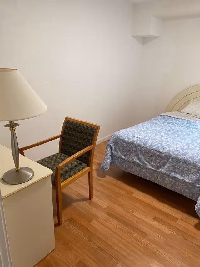 A simple bedroom with wooden flooring, a bed with a blue floral bedspread, a wooden chair with patterned upholstery, and a small white nightstand with a lamp on it. The walls are plain white.