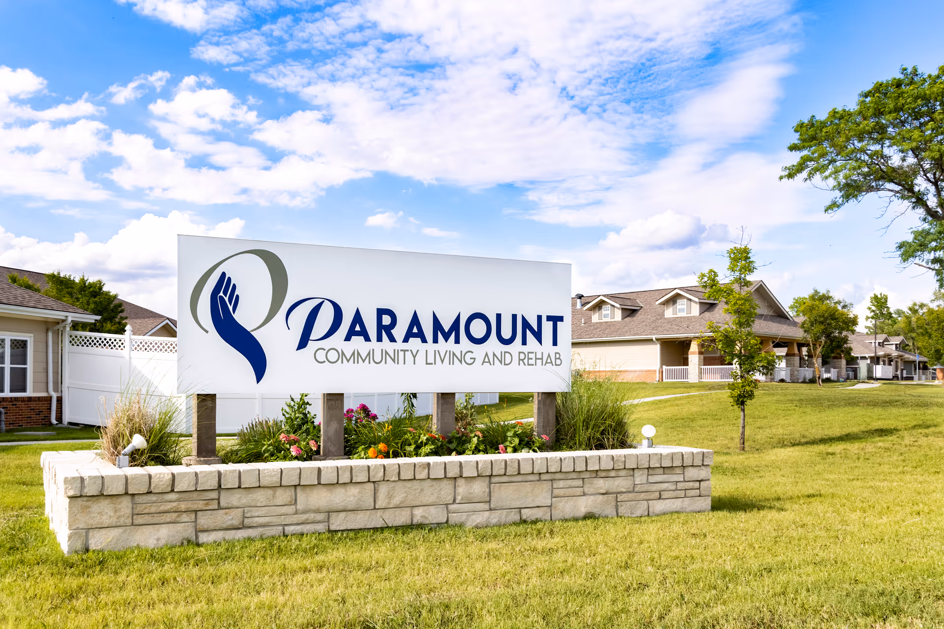 Outdoor view of Paramount Community Living and Rehab sign with a landscaped stone base, green grass, trees, and residential-style buildings in the background under a partly cloudy blue sky.