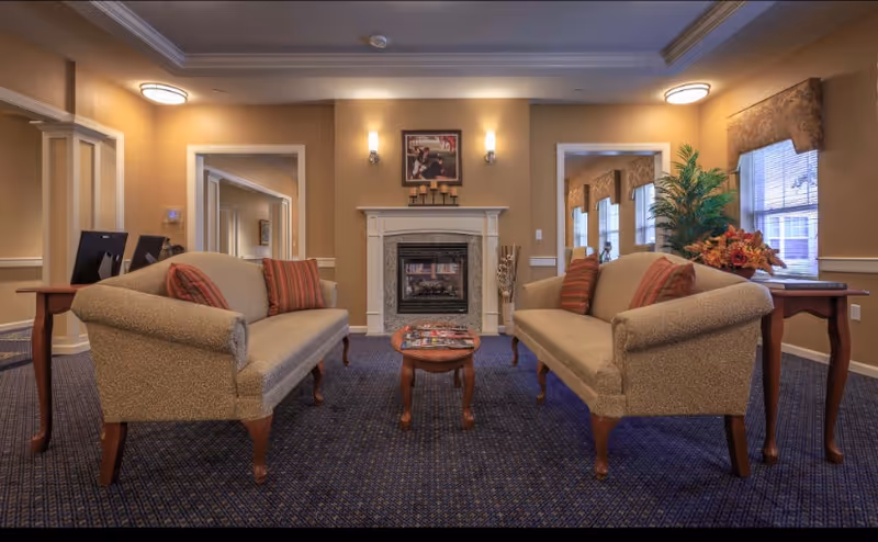 A cozy living room area with two beige upholstered sofas facing each other across a small wooden coffee table. The room features a carpeted floor, a fireplace centered on the far wall with a framed picture and candles above it, wall sconces on either side of the fireplace, and windows with valances letting in natural light. There are side tables next to each sofa, one with a computer monitor and the other with a floral arrangement and a potted plant.