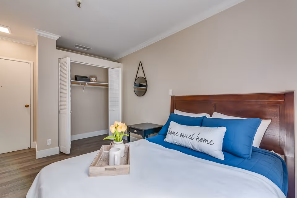 A cozy bedroom with a wooden bed frame, blue bedding, and a white pillow with the words 'home sweet home'. There is a small black nightstand with a round mirror hanging above it. An open closet with white louvered doors is visible, along with a wooden floor and beige walls.