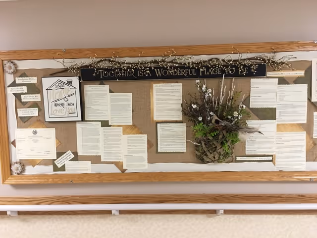 A bulletin board with various printed notices and papers pinned on it, decorated with a floral arrangement and a sign at the top that reads 'Together is a Wonderful Place to Be'. The board is framed with wood and mounted on a wall above a handrail.