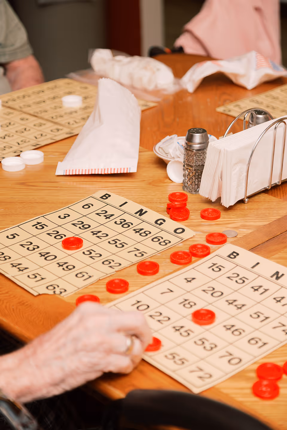 Close-up view of a wooden table with bingo cards and red bingo chips scattered on them. A person's hand is placing a chip on one of the cards. There are also napkins in a holder, a salt shaker, and some rolled-up napkins on the table.