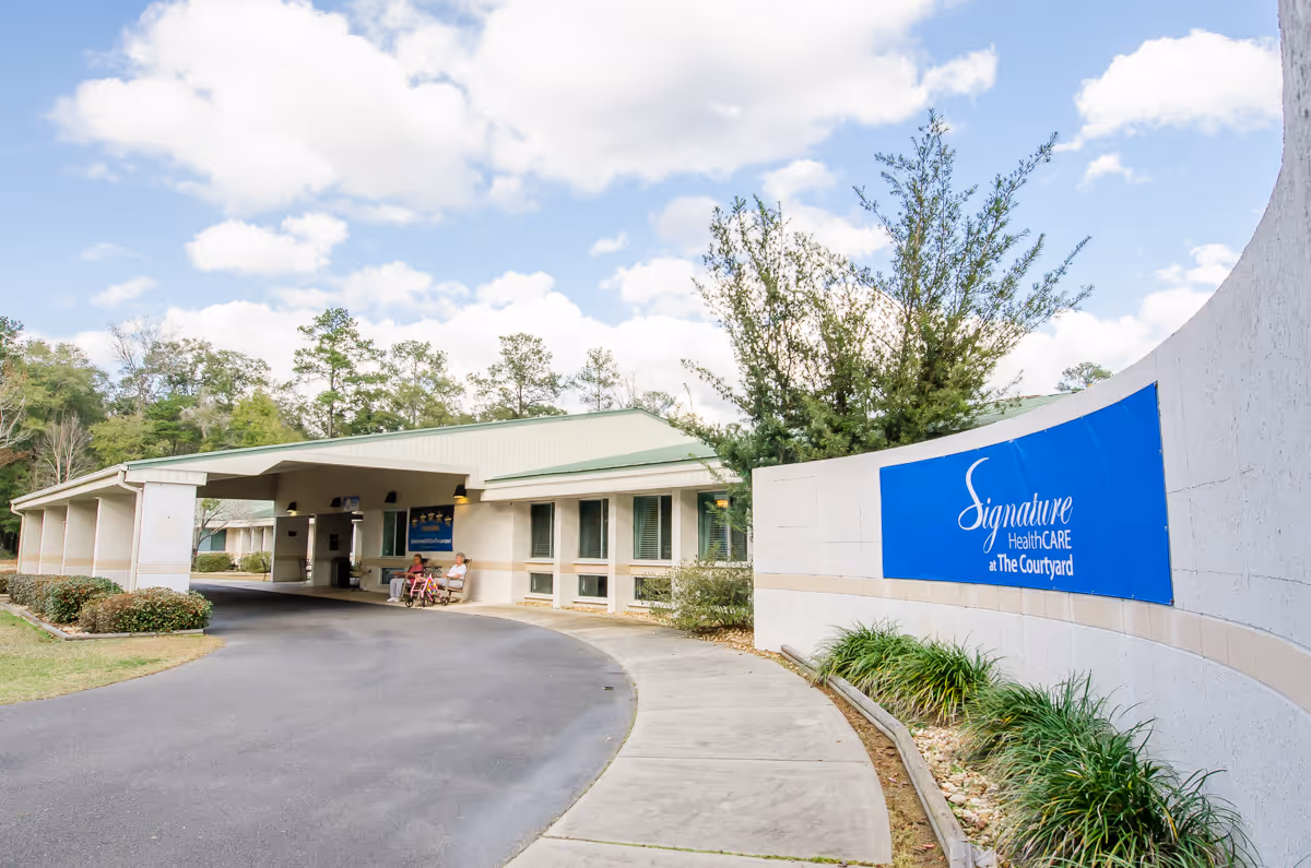 Front exterior and entrance drive of a senior healthcare facility with a blue 'Signature HealthCARE at The Courtyard' sign and covered drop-off area.