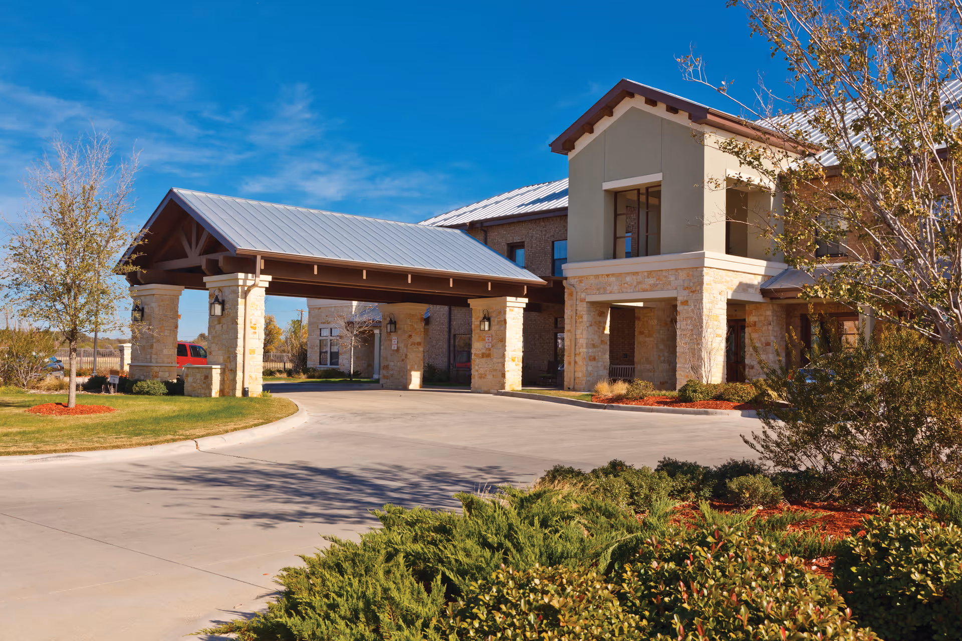 Front entrance of a senior living building with a covered porte-cochère, stone façade and landscaped driveway under a blue sky.
