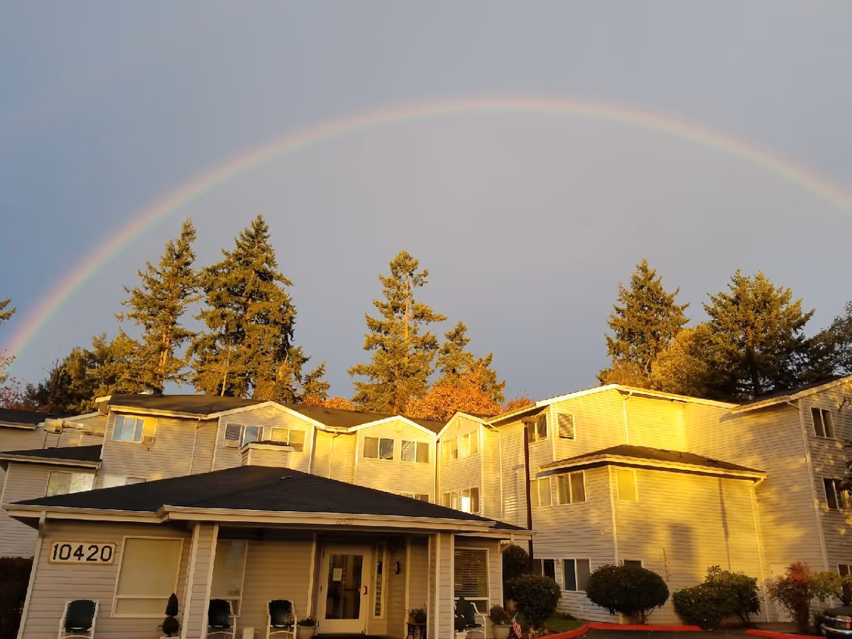 Front exterior of a senior living building (10420) with trees and a rainbow arching overhead.