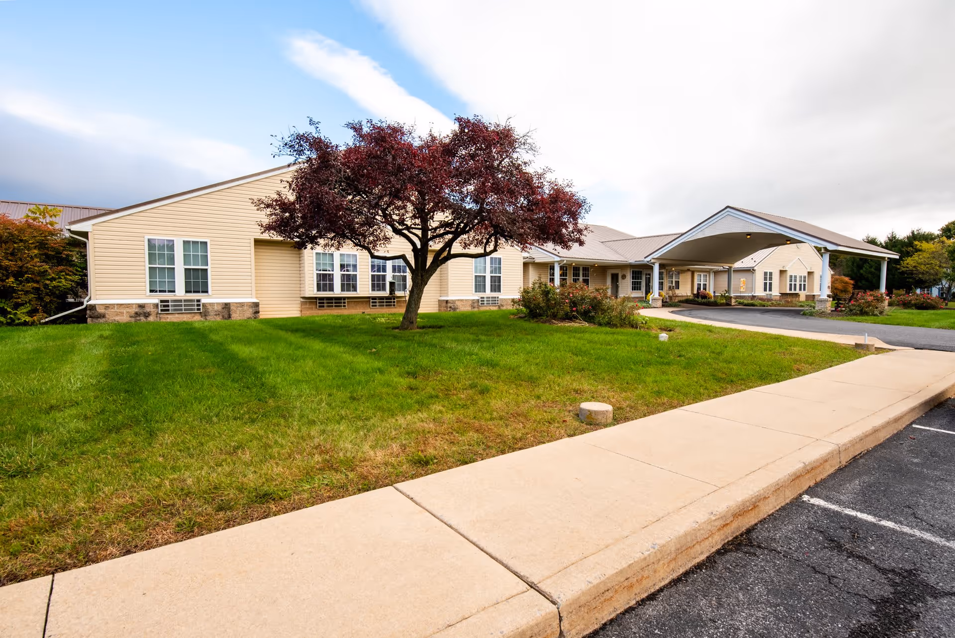Exterior view of Wynwood House, a single-story building with beige siding and multiple windows. There is a tree with dark red leaves in the grassy front yard and a covered entrance driveway. The sky is partly cloudy.