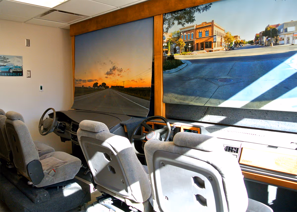 Interior view of a driving simulator setup with two steering wheels and multiple seats facing large screens displaying road scenes, one showing a sunset on a rural road and the other a sunny town intersection.