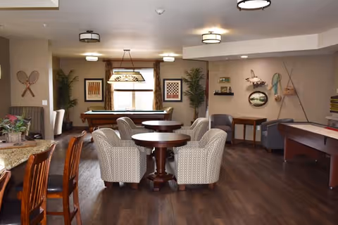 Community recreation room with round tables and upholstered chairs, a pool table, shuffleboard table, and additional seating and decor.