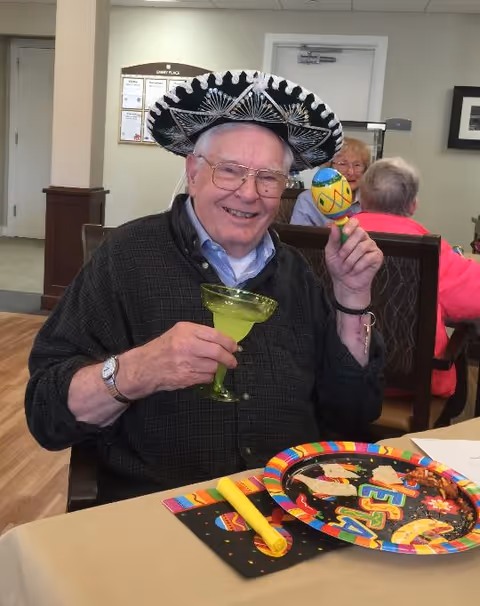 An elderly man wearing glasses and a black and white sombrero hat is smiling while holding a green margarita glass in one hand and a colorful maraca in the other. He is seated at a table with festive decorations including a colorful plate and a yellow party blower. In the background, two other elderly people are seated and engaged in conversation in a communal dining area.