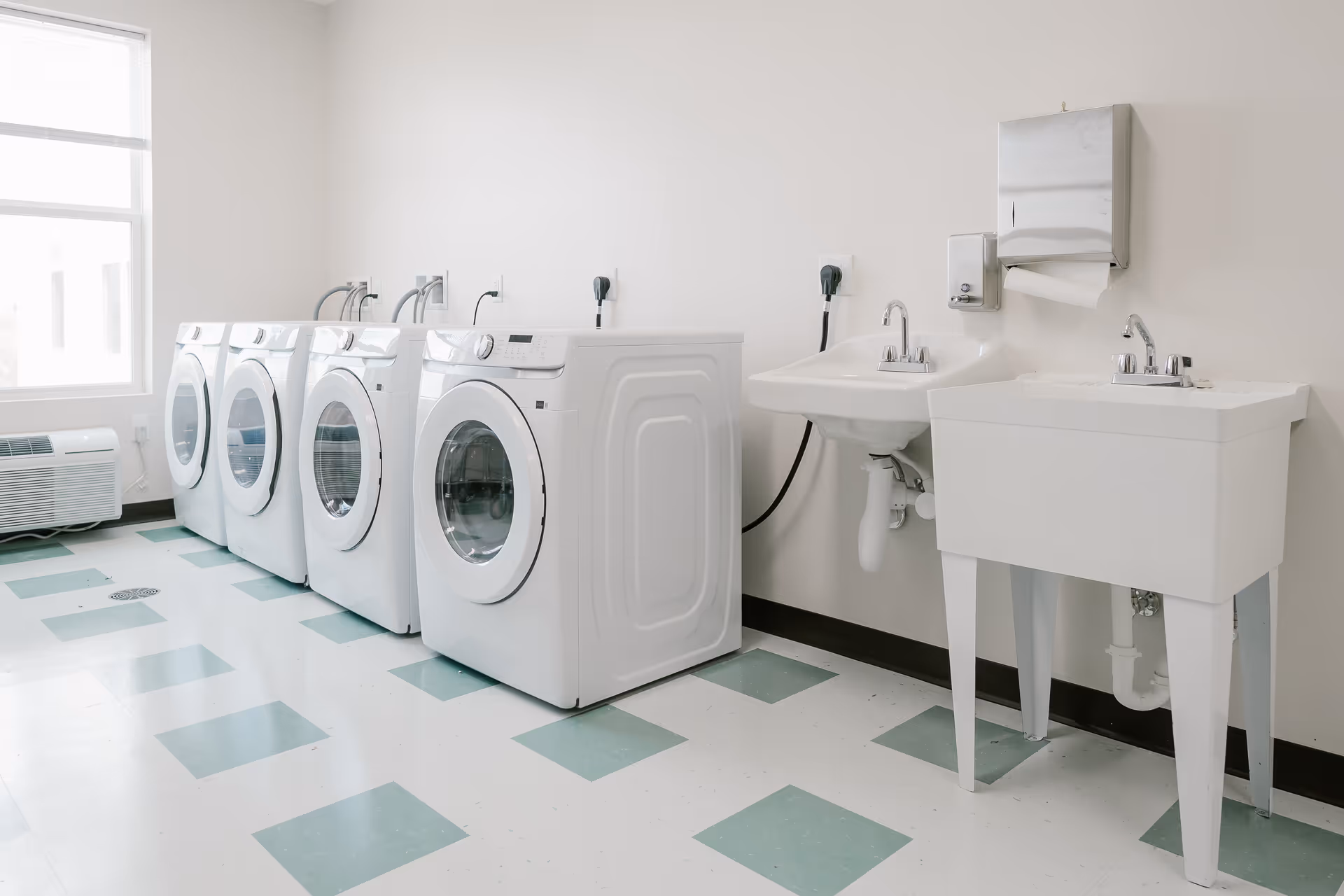 A clean laundry room with four front-loading washing machines lined up against a wall. To the right, there are two sinks, one wall-mounted and one utility sink with legs. The floor has a white and green checkered pattern, and there is a window on the left side letting in natural light.