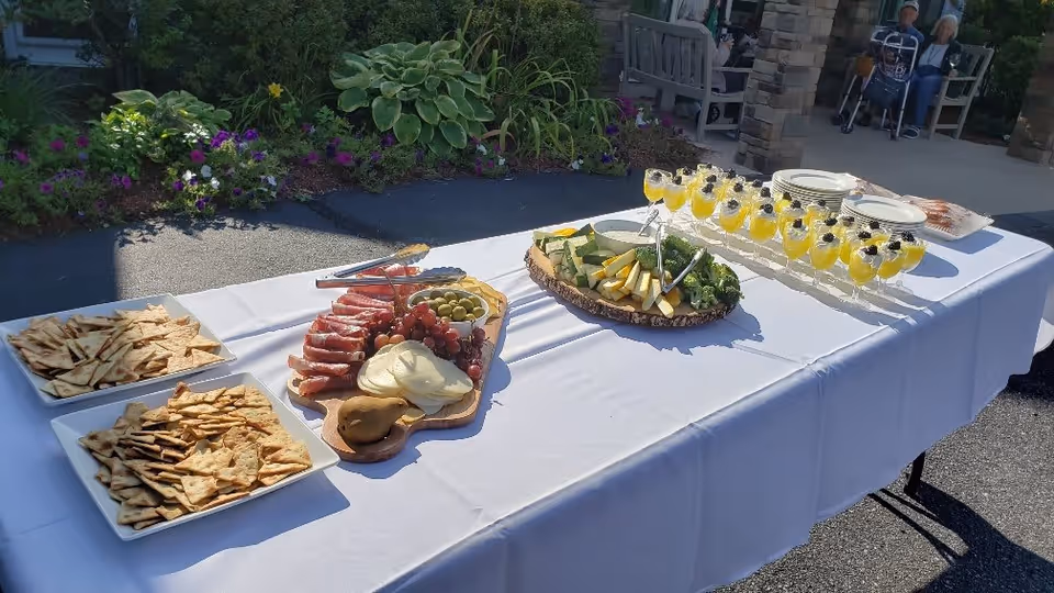 Outdoor buffet table covered with a white tablecloth displaying plates of crackers, a charcuterie board with meats, cheeses, grapes, and olives, a platter of sliced vegetables with dip, and glasses filled with a yellow beverage garnished with berries. In the background, there are plants, stone pillars, and two elderly people sitting on benches.