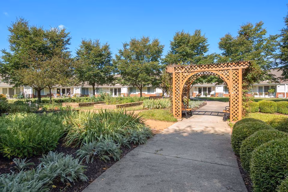 A landscaped garden area with a concrete pathway leading through a wooden lattice archway. The garden features various green plants, shrubs, and trees. In the background, there is a single-story building with multiple doors and windows, likely part of a senior living facility.