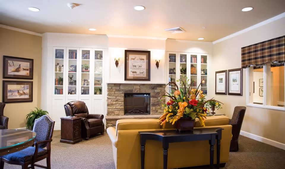 A cozy living room area in an assisted living facility featuring a stone fireplace with a framed animal print above it, flanked by white built-in cabinets with glass doors displaying books and decorative items. The room has a tan sofa with a black console table behind it holding a large floral arrangement, a brown leather armchair, and several upholstered chairs around a glass-top table. The walls are adorned with framed artwork and a window with a plaid valance.