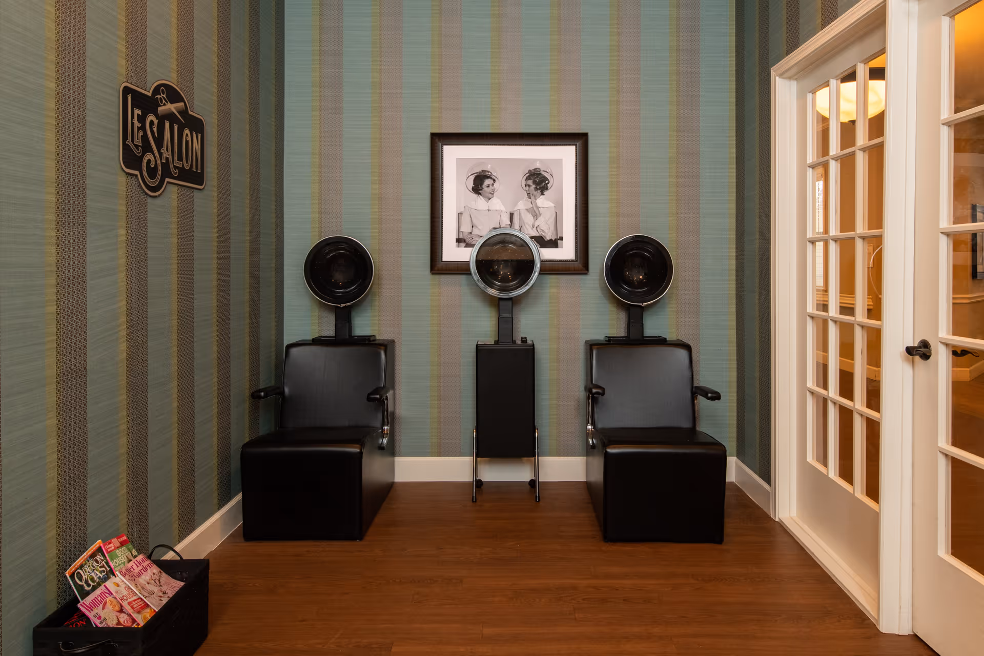 Interior view of a salon area with two black salon chairs equipped with hair dryers. The walls have vertical striped wallpaper in shades of green and beige. A framed black and white photo of two women under hair dryers hangs on the wall between the chairs. To the left, a sign reads 'Salon' and a basket with magazines is on the floor. To the right, there is a white-framed glass door.