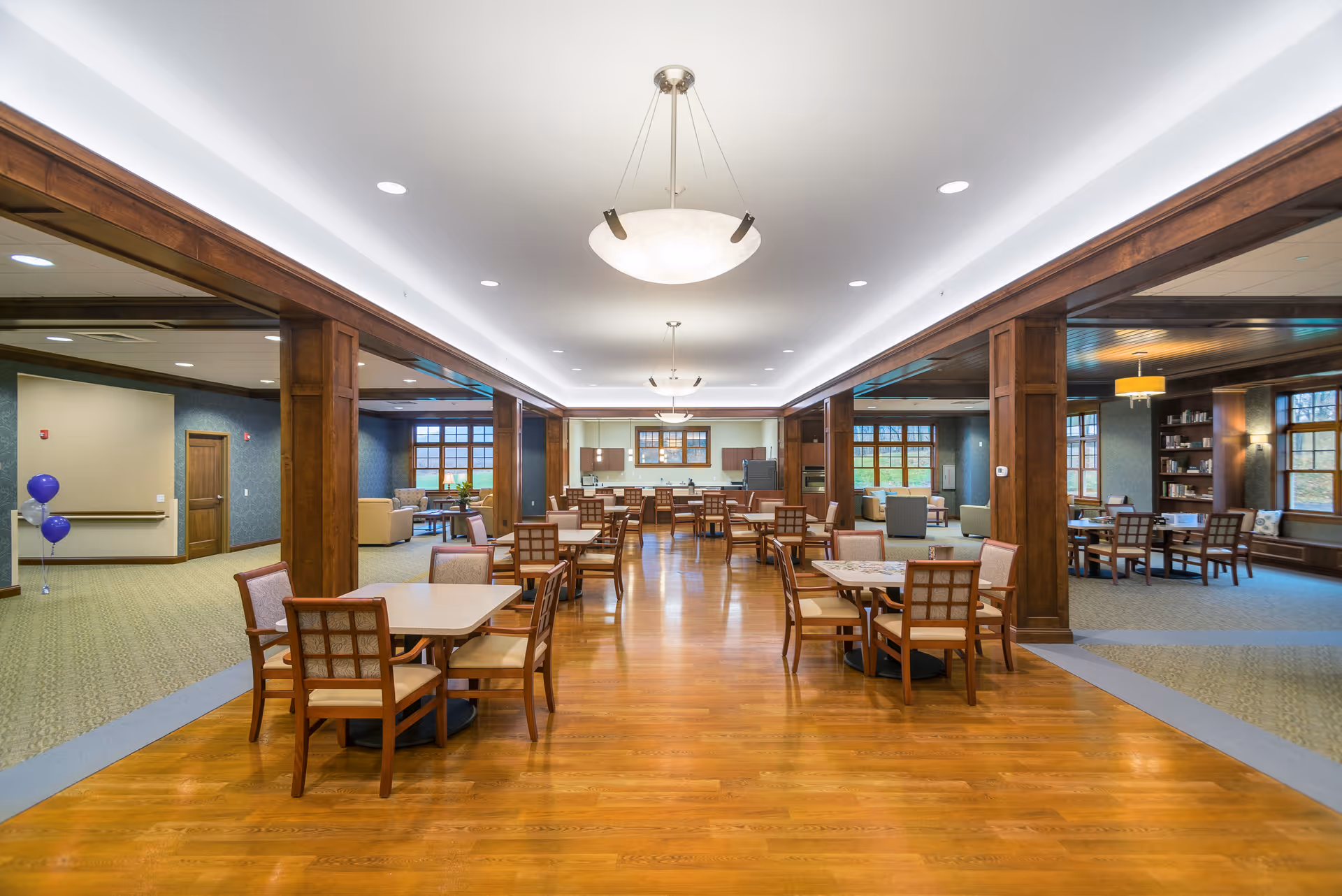 Spacious dining area in Armstrong Memory Care Assisted Living with wooden floors, multiple square tables each surrounded by four chairs, and large windows letting in natural light. The room features wooden pillars and trim, modern ceiling lights, and adjacent lounge areas with comfortable seating and bookshelves.