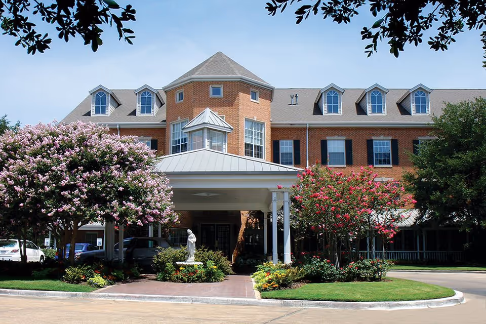 Front exterior view of Caruth Haven Court, a large brick building with multiple windows and dormers. The entrance features a covered driveway with white columns, surrounded by well-maintained landscaping including flowering trees and bushes. A statue is visible near the entrance.