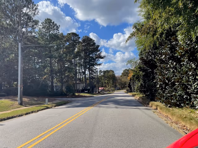 A quiet tree-lined residential street with houses and a partly cloudy blue sky.