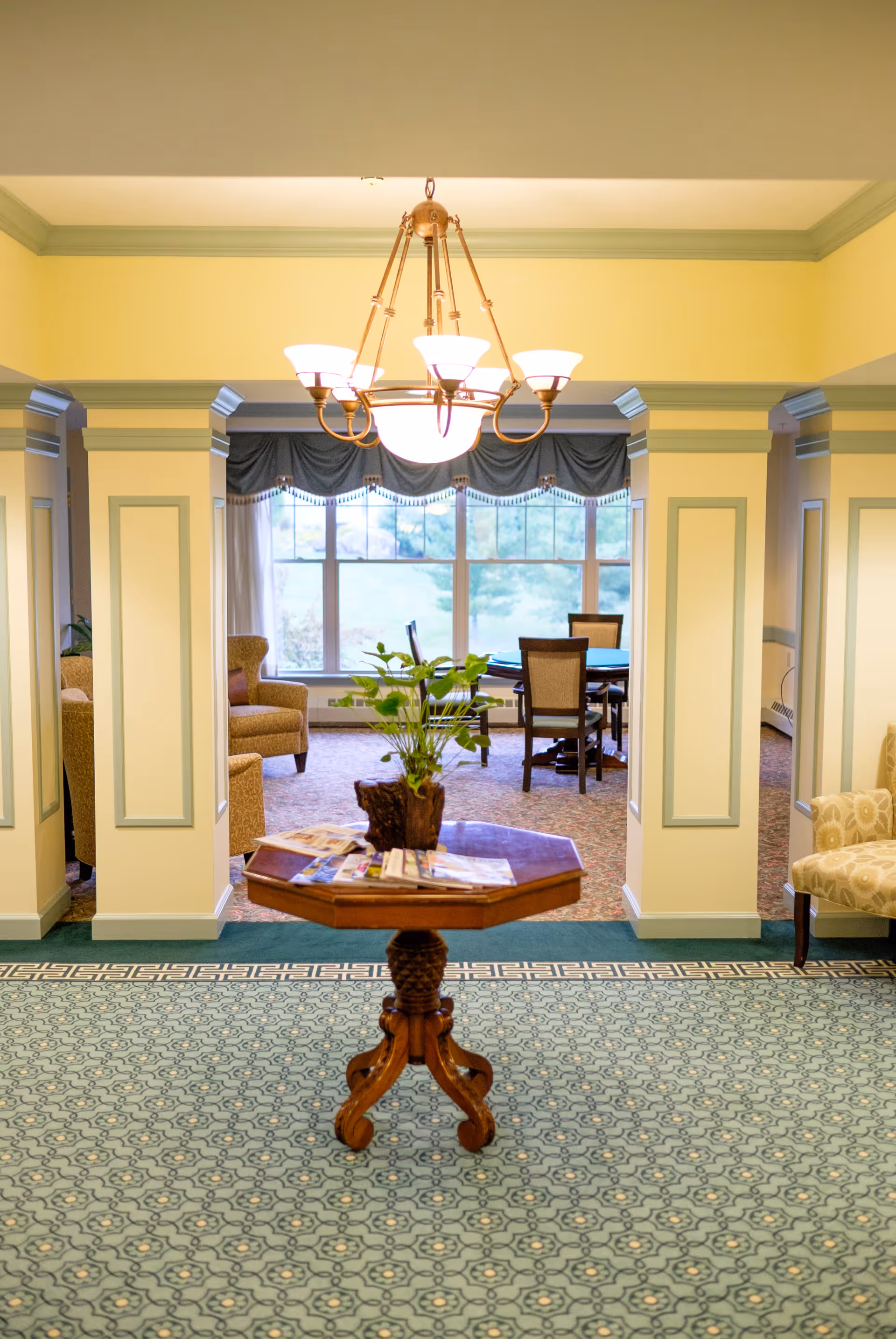 Interior view of a senior living facility showing a small wooden table with a plant and magazines in the foreground, a chandelier hanging from the ceiling, and a sitting area with chairs and a table near large windows in the background.