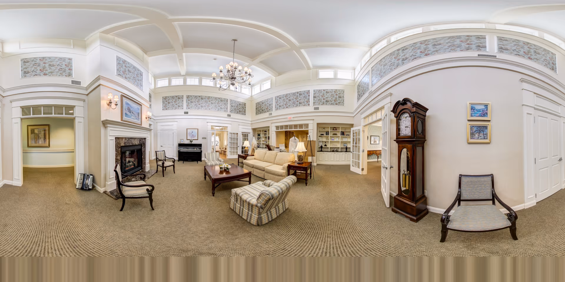 A spacious and elegantly decorated living room with high ceilings and large windows near the ceiling. The room features a beige sofa, a plaid armchair, a wooden coffee table, and several chairs arranged around a fireplace with a framed painting above it. There is a grandfather clock against one wall and built-in shelves with books and decorative items. The walls have floral wallpaper accents near the ceiling, and a chandelier hangs from the ceiling.