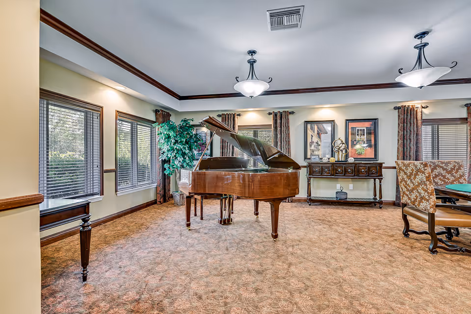 A spacious, well-lit room with a grand piano in the center, surrounded by large windows with blinds and curtains. The room features a patterned carpet, a wooden sideboard with decorative items and framed pictures on the wall above it, and a table with upholstered chairs on the right side. The ceiling has recessed lighting and two hanging light fixtures.