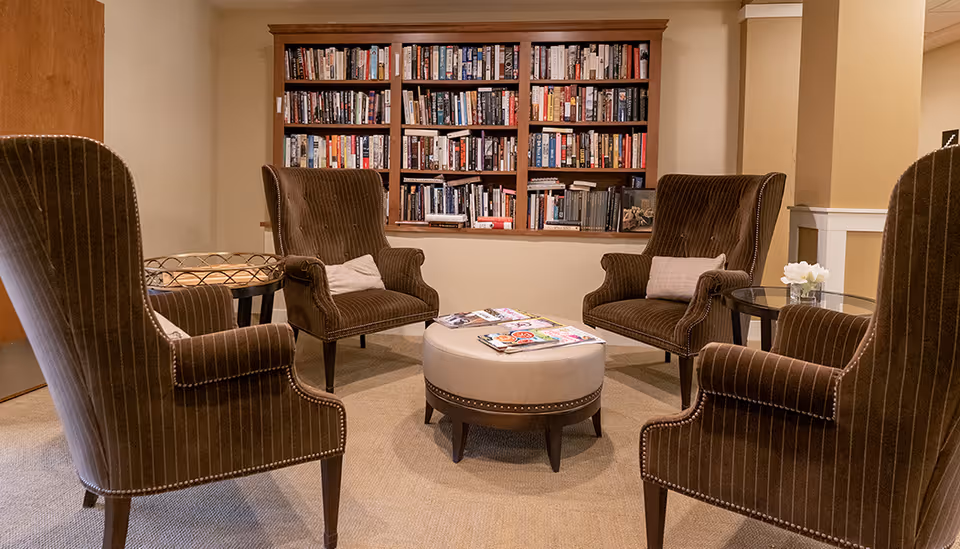 Cozy seating area with four upholstered armchairs arranged around an ottoman and a bookshelf filled with books.