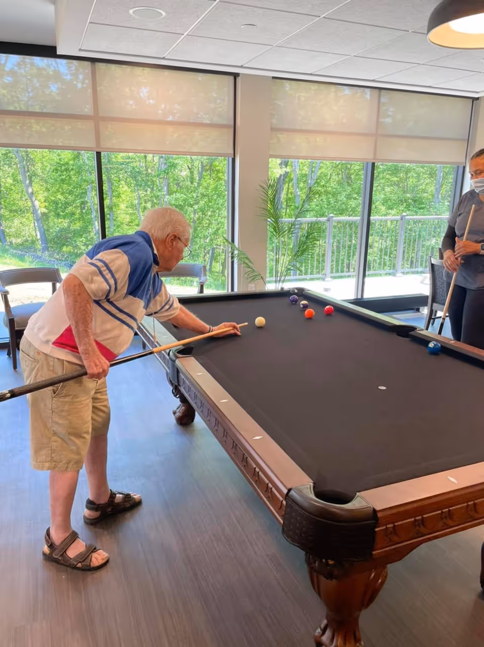 An elderly man playing pool indoors, aiming a cue stick at the white ball on a black felt pool table. Another person wearing a mask stands nearby holding a cue stick. Large windows in the background show green trees and a balcony outside.