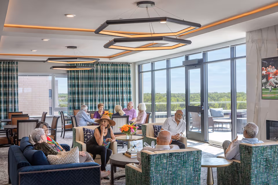 A bright and spacious senior living common area with large windows showing a green outdoor view. Several elderly people are seated on sofas and chairs, engaged in conversation. The room features modern hexagonal ceiling lights, patterned curtains, a fireplace with a mounted TV showing a football game, and a round table with a flower arrangement.