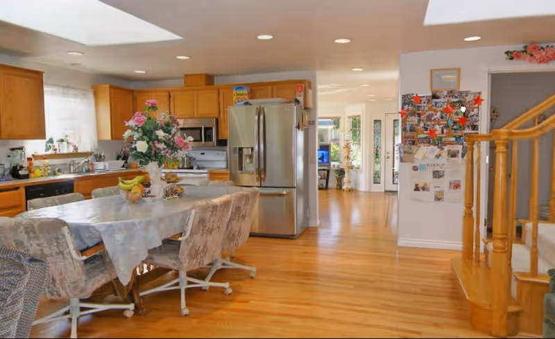 A spacious kitchen and dining area with wooden floors and cabinetry. The kitchen features a stainless steel refrigerator, microwave, and stove. A dining table covered with a floral tablecloth is surrounded by cushioned chairs on casters. A vase with flowers and a bowl of fruit are on the table. In the background, there is a staircase with wooden railings and a wall covered with photos and notes. The area is well-lit with recessed ceiling lights and natural light from windows.
