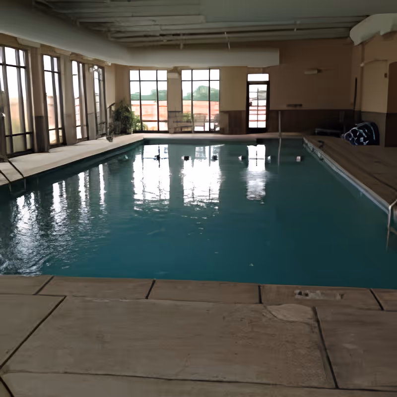 Indoor swimming pool with clear blue water surrounded by large windows letting in natural light. The pool area has a tiled floor and some pool equipment is visible on the right side.