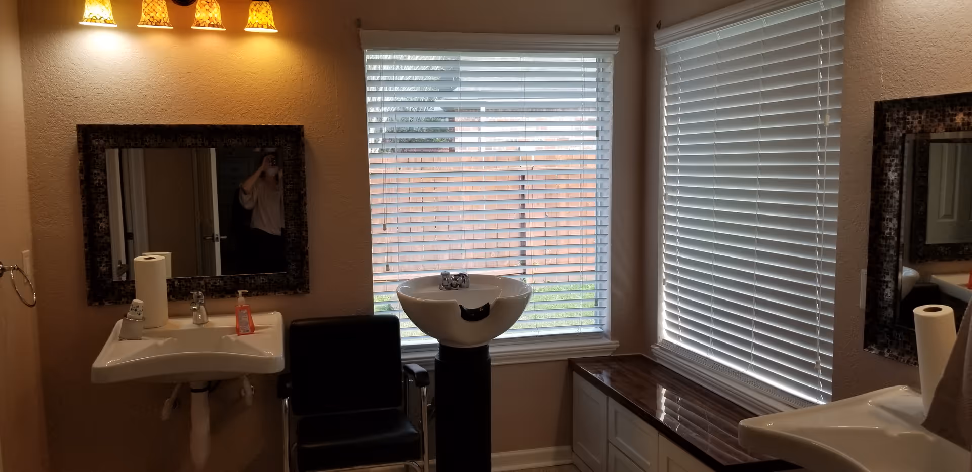 Interior view of a room with two sinks, two mirrors, a black chair, and a white basin on a stand. There are two windows with white blinds letting in natural light. The walls are beige, and there are three yellowish wall lights above one of the mirrors.