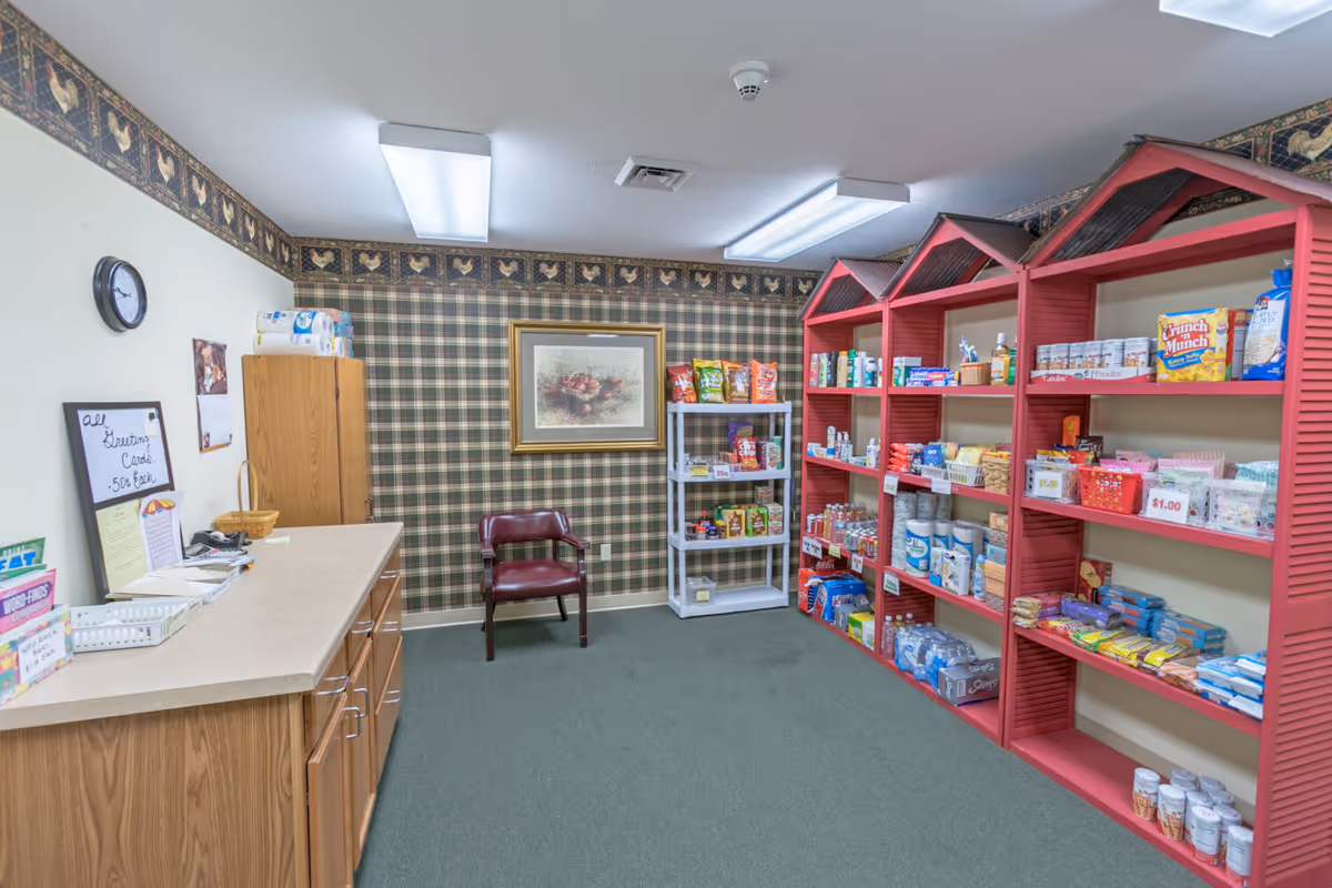 Interior room with green carpet and plaid wallpaper featuring a rooster border. On the right side, red shelves shaped like small houses hold various snacks, toiletries, and household items. A small white shelf also holds snacks. On the left side, a wooden counter with drawers and cabinets has a sign advertising greeting cards for 50 cents each. A maroon chair is placed against the back wall beneath a framed picture of fruit. The ceiling has fluorescent lights.