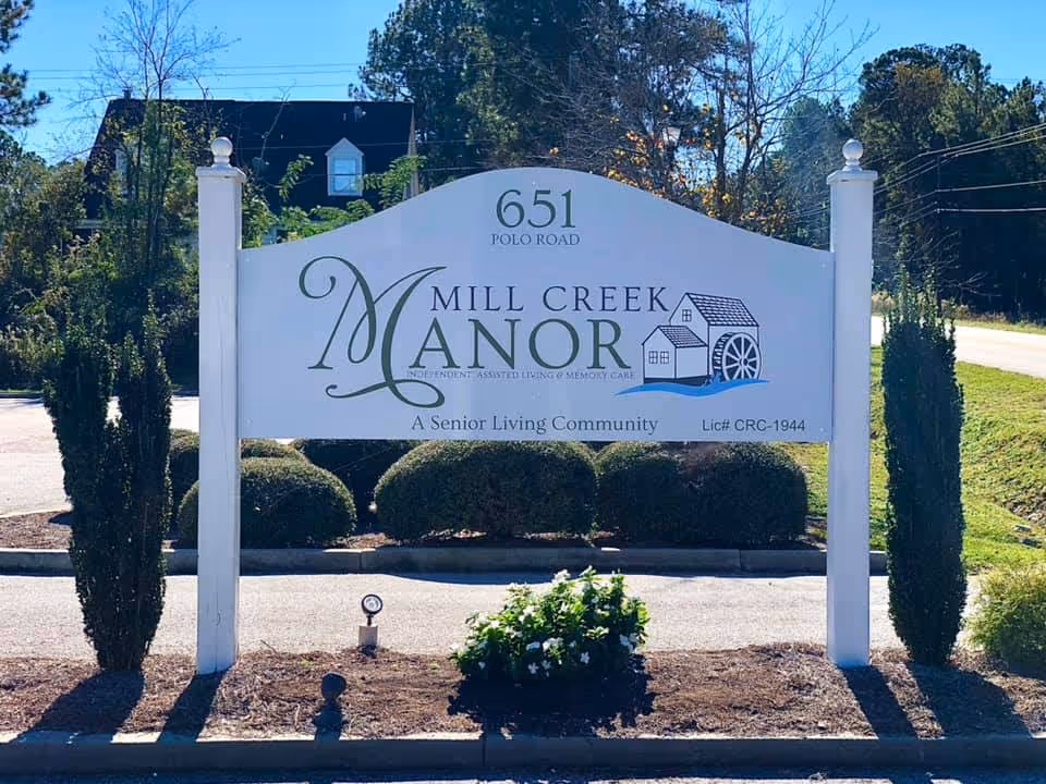 Outdoor sign for Mill Creek Manor Senior Living Community located at 651 Polo Road, surrounded by trimmed bushes and trees with a clear blue sky in the background.