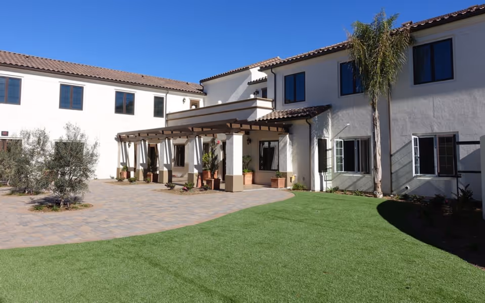 Exterior view of a senior living facility building with white walls and a tiled roof under a clear blue sky. The building has multiple windows, a covered entrance with wooden beams, potted plants, a paved walkway, and a green lawn with a palm tree.