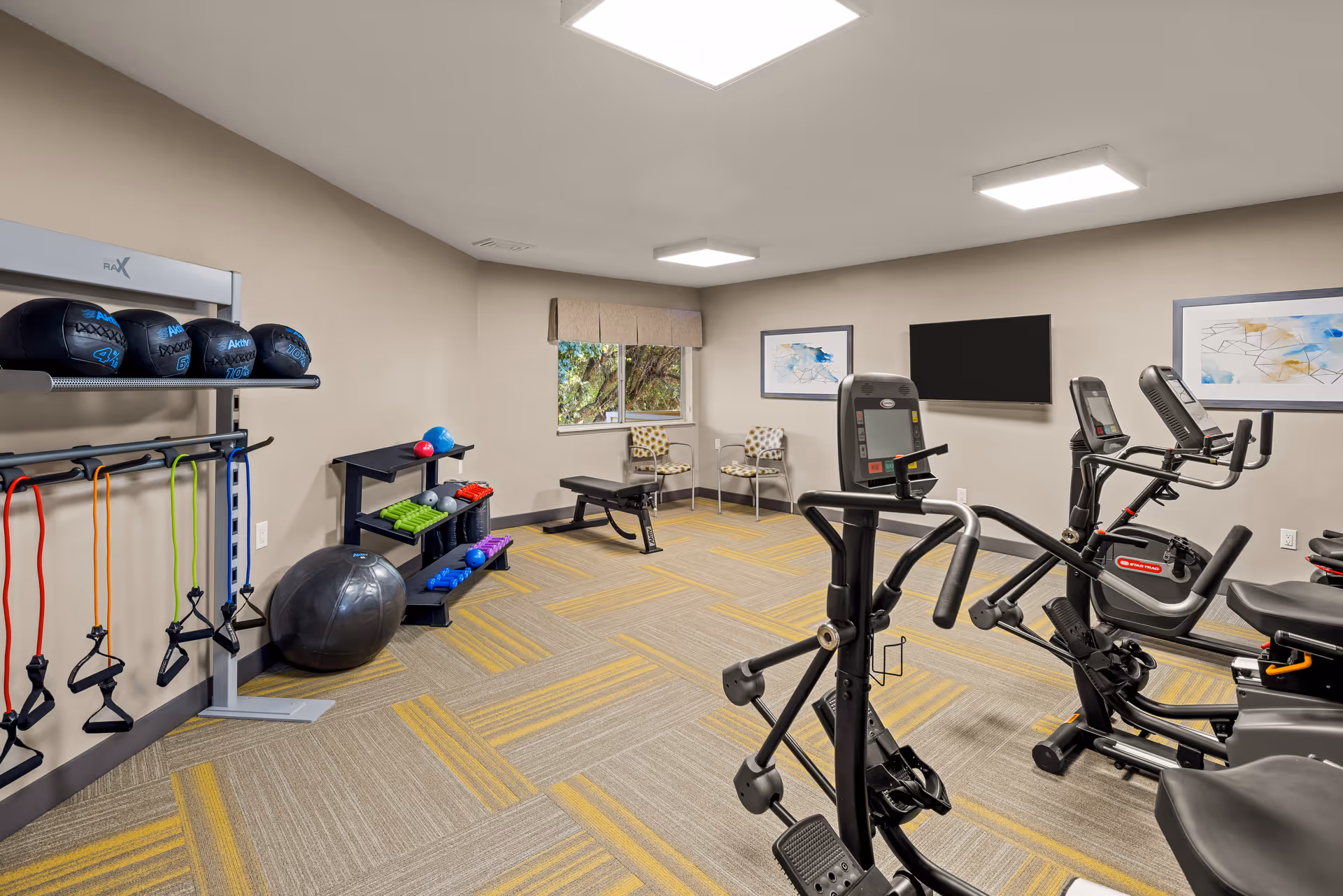 A senior living community fitness room with exercise equipment including stationary bikes, resistance bands, medicine balls, and a stability ball. The room has beige walls, patterned carpet with yellow accents, two chairs near a window, and framed artwork on the walls.