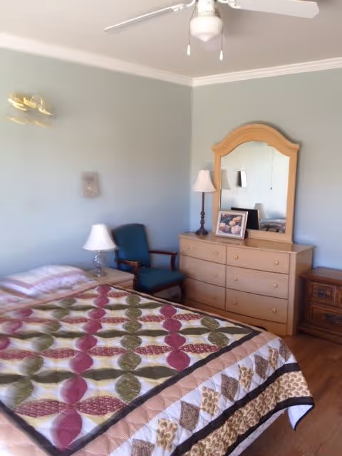 A bedroom with a bed covered by a colorful quilt, a wooden dresser with a mirror, a table lamp, a framed photo, a wooden nightstand, and a green upholstered chair. The walls are painted light blue and there is a ceiling fan above.