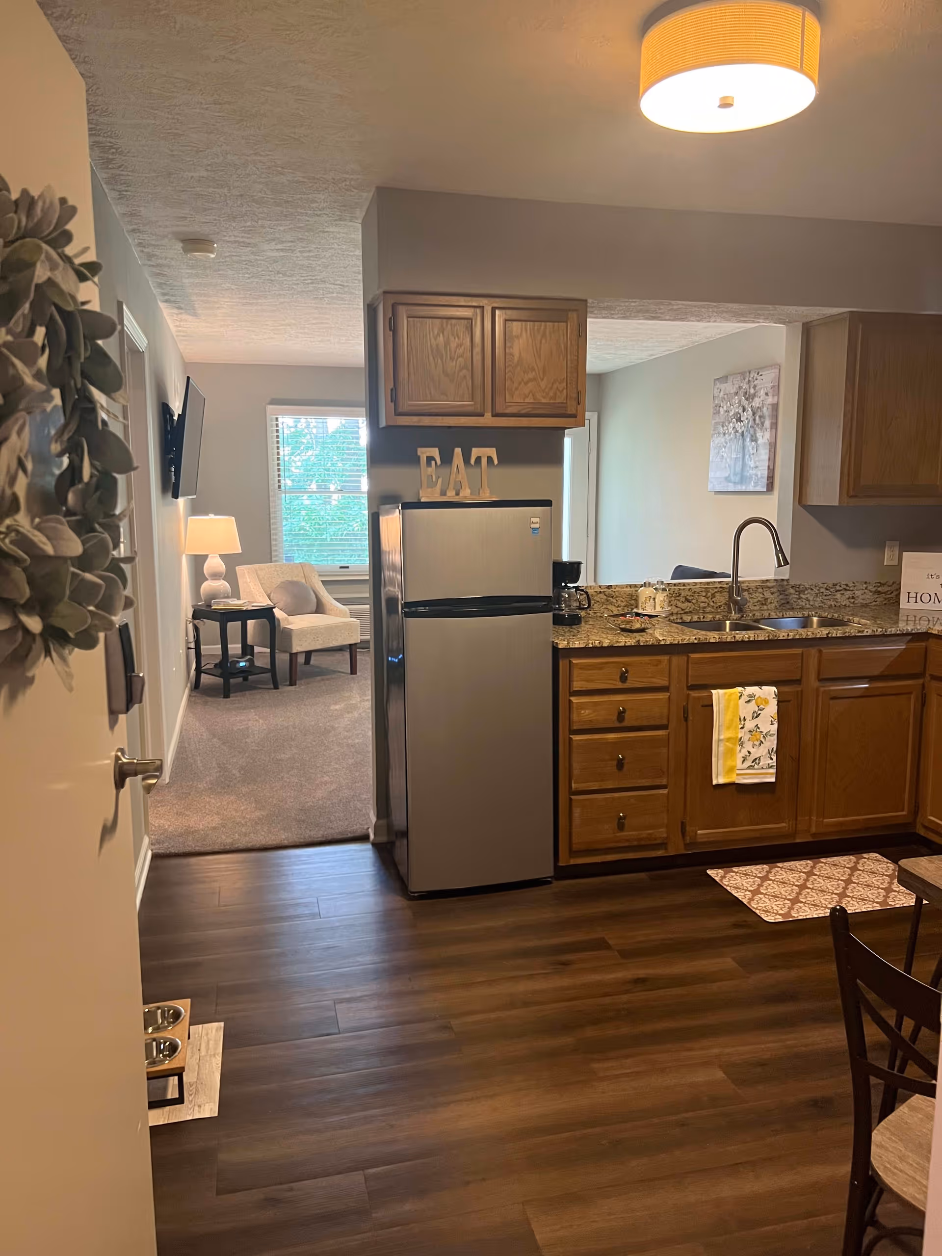 View of a kitchen area with wooden cabinets, a stainless steel refrigerator, granite countertops, and a sink. A decorative sign spelling 'EAT' is placed on top of the refrigerator. In the background, there is a living area with a window, a lamp on a side table, and an upholstered chair. The floor is dark wood, and a ceiling light fixture is visible.