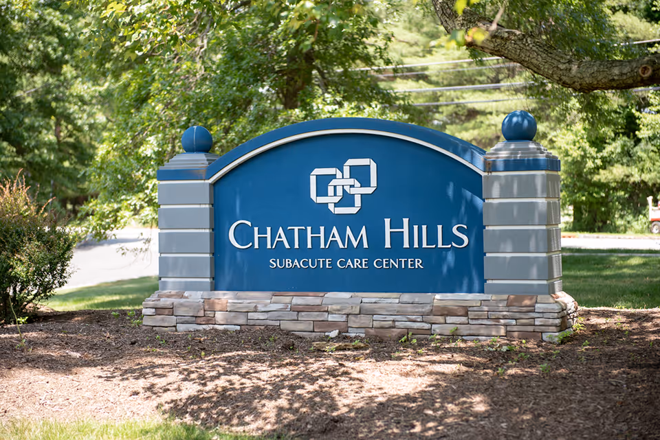 Blue stone entrance sign reading "Chatham Hills Subacute Care Center" set in landscaped grounds with trees in the background.