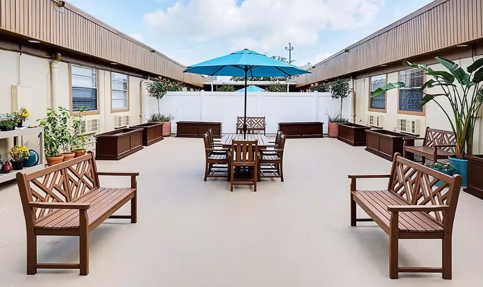 Outdoor courtyard area with wooden benches and a wooden table with chairs under a blue umbrella. The space is surrounded by a building with windows and potted plants along the walls.