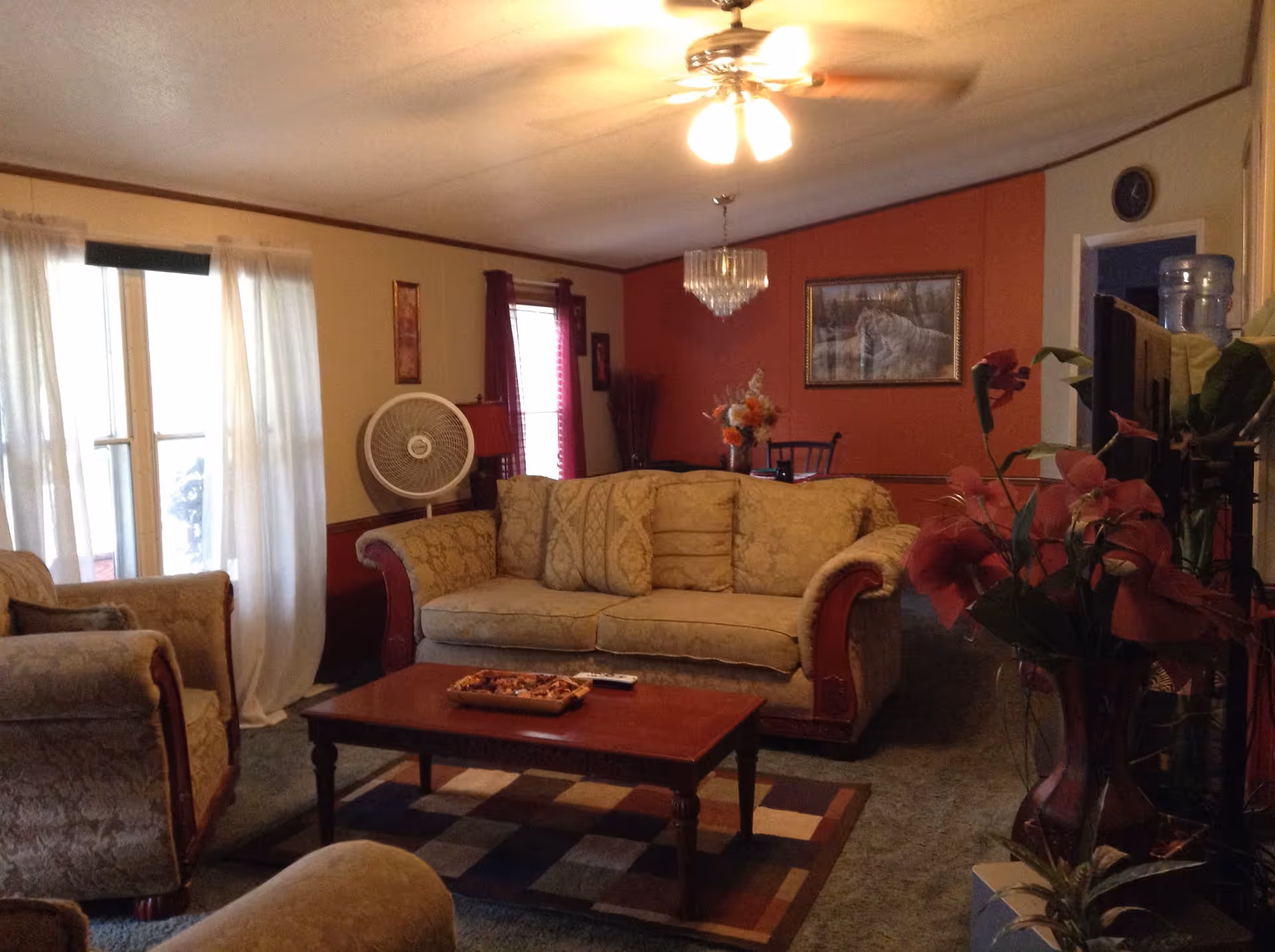 A cozy living room with a beige patterned sofa and matching armchairs arranged around a wooden coffee table on a multicolored rug. The room has a ceiling fan with lights, a large window with sheer white curtains, a standing fan, and a dining area with a chandelier and a framed picture on a red accent wall in the background. There are also decorative plants and a water dispenser visible.