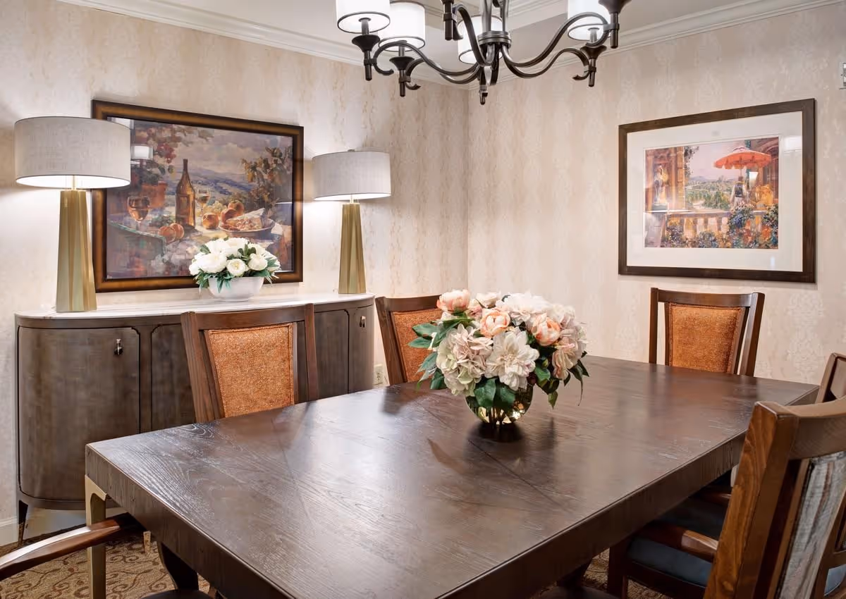 A formal dining room featuring a dark wooden table with a floral centerpiece, upholstered chairs, a sideboard with lamps, and framed artwork on patterned walls.