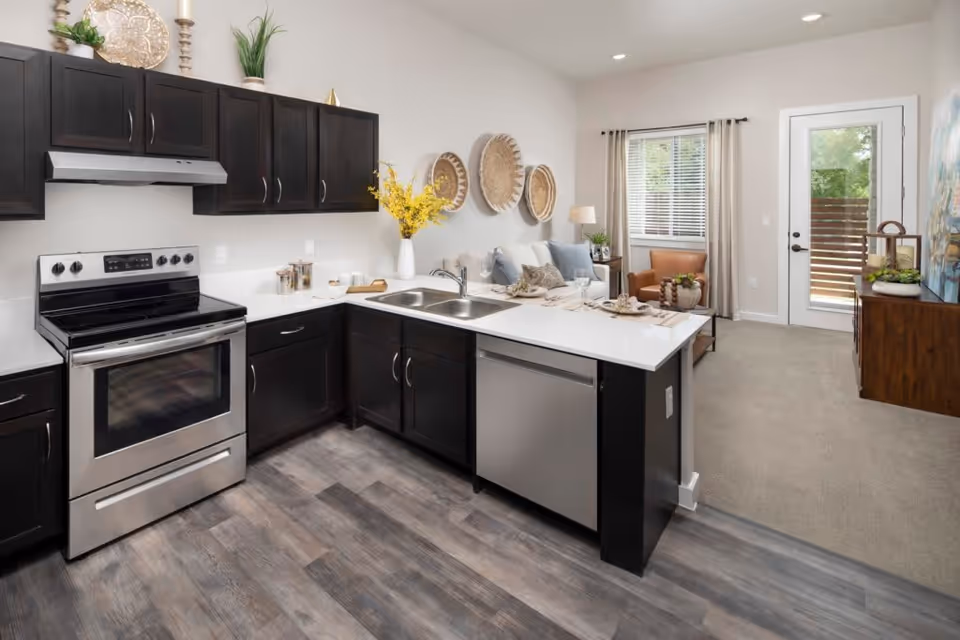 Modern kitchen with dark wood cabinets, stainless steel stove and dishwasher, white countertops, and a small living area with a sofa, armchair, window with blinds, and a door leading outside. Decorative baskets hang on the wall above the sofa.