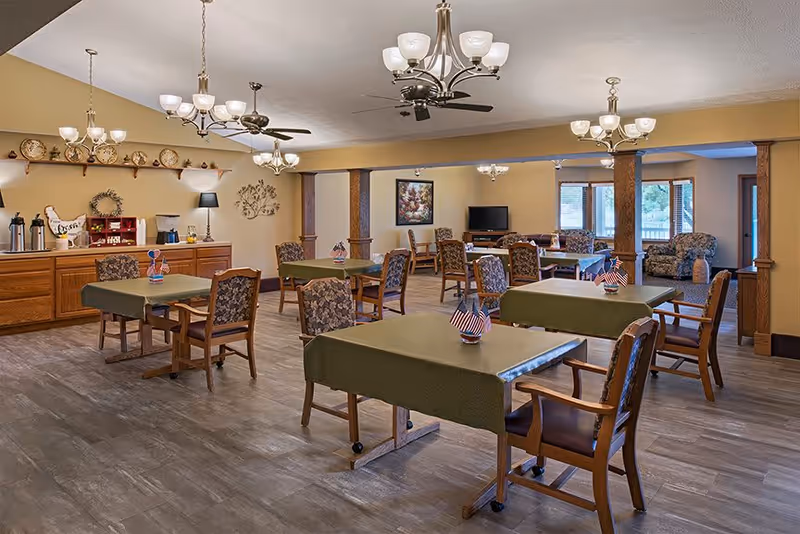 A spacious dining room with several tables covered in green tablecloths, each table decorated with small American flag centerpieces. The room features wooden chairs with floral upholstery, wooden columns, and multiple chandeliers and ceiling fans providing lighting. A sideboard with coffee and tea supplies is against one wall, and a seating area with armchairs and a TV is visible in the background near large windows.