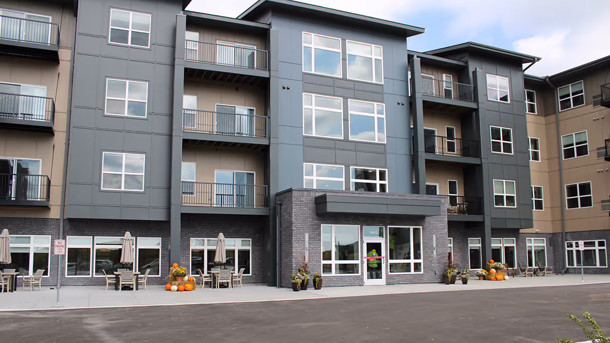 Front exterior view of a modern multi-story senior living facility with balconies, large windows, and a main entrance. There are outdoor seating areas with tables, chairs, and umbrellas, along with seasonal decorations including pumpkins and flowers near the entrance.