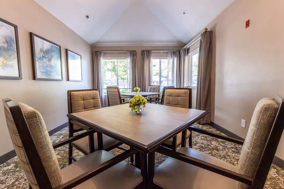 A bright dining room with two square wooden tables, each surrounded by four cushioned chairs. The room has large windows with beige curtains allowing natural light to fill the space. There are three framed abstract paintings on one wall and small floral centerpieces on each table. The carpet has a patterned design in neutral tones.