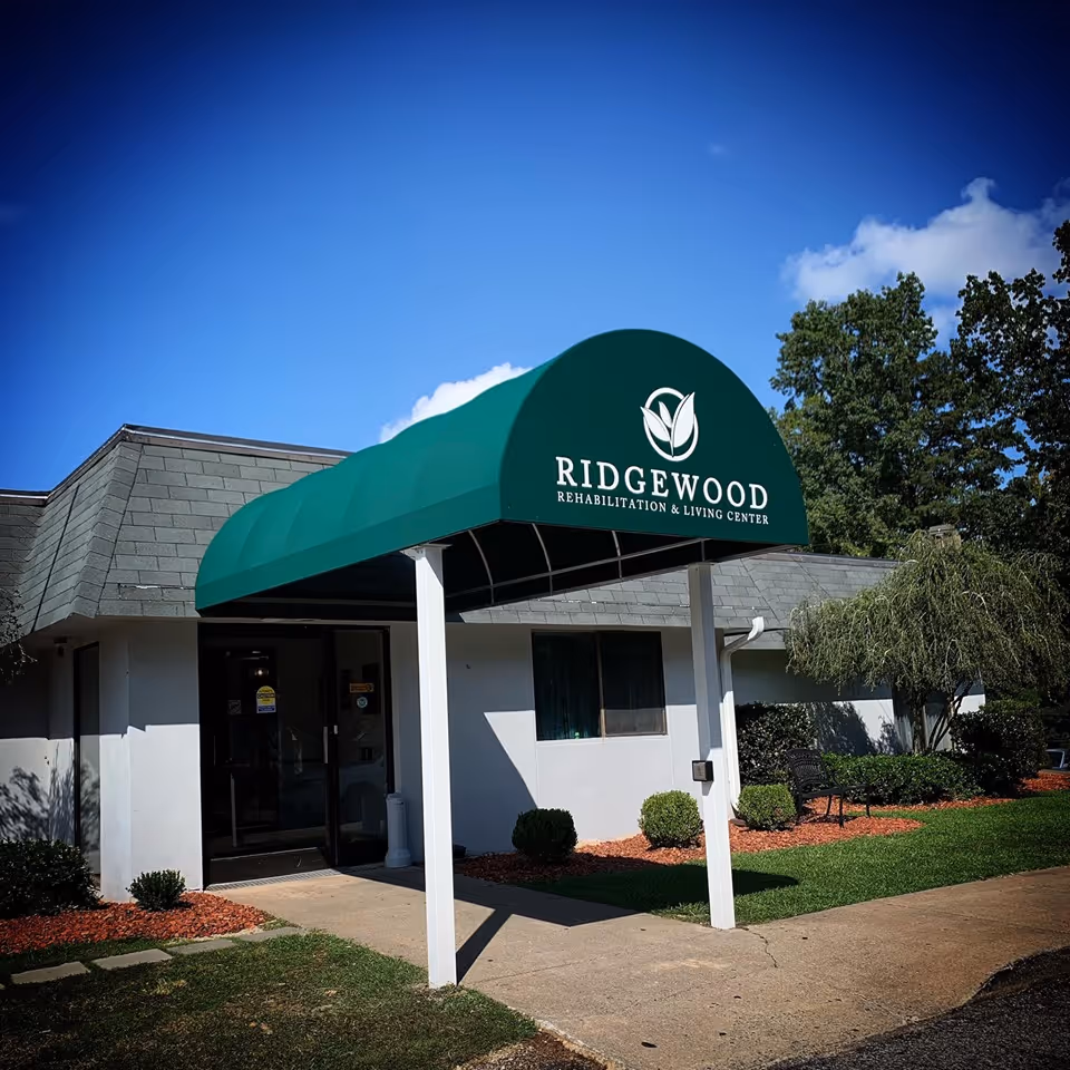 Exterior view of Ridgewood Rehabilitation & Living Center showing the entrance with a green awning displaying the facility's name and logo. The building is white with a gray roof, surrounded by neatly trimmed bushes and a small tree under a clear blue sky.