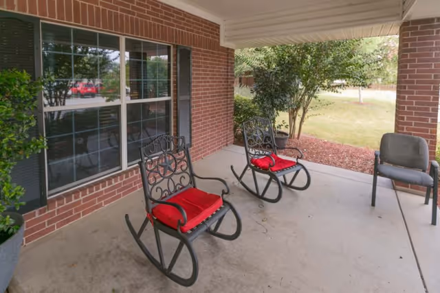 Covered front porch with two wrought-iron rocking chairs with red cushions beside a brick wall and window overlooking a lawn.