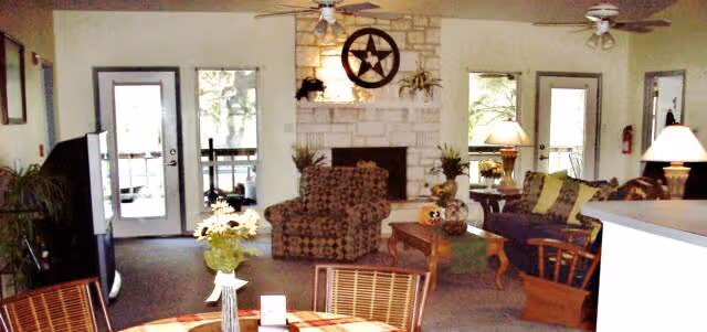 Sunlit communal living room with sofas, an armchair, a stone fireplace, ceiling fans, and a dining table near glass doors and windows.